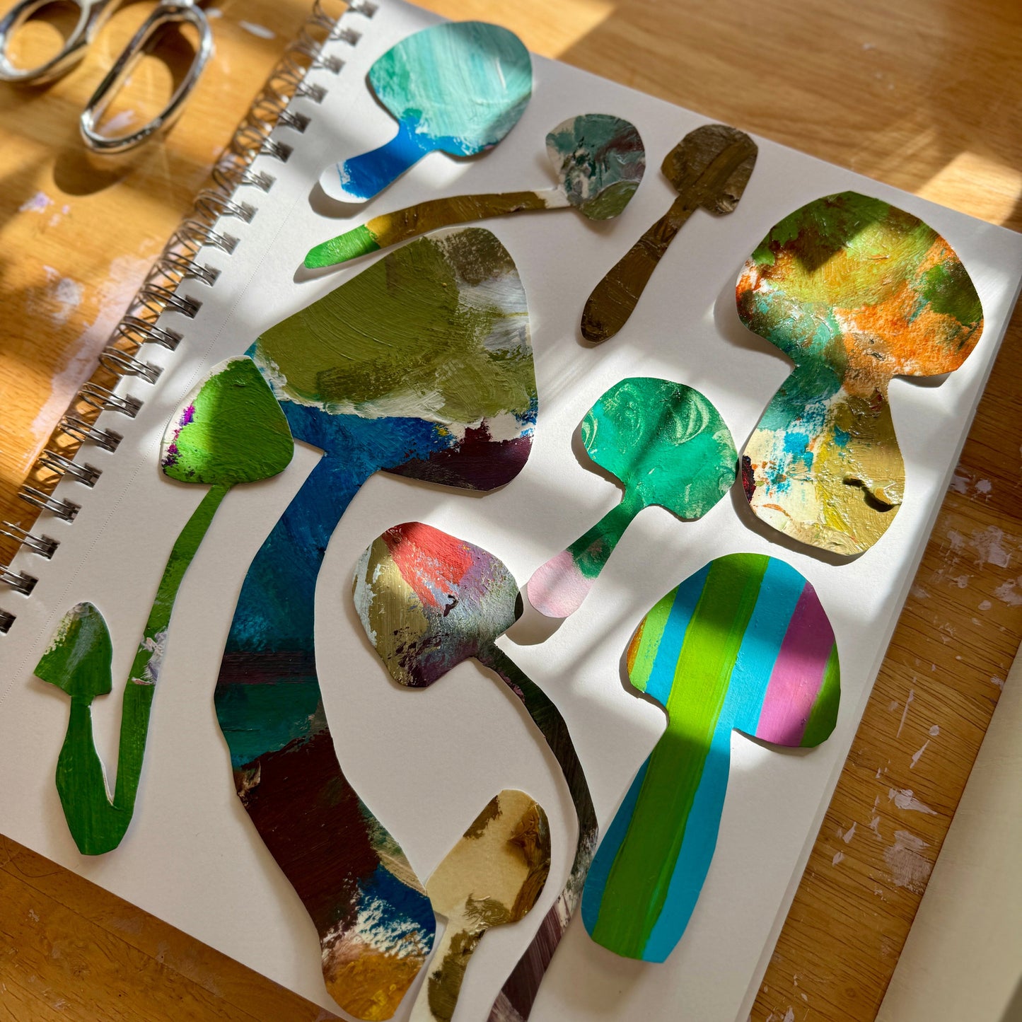Large and small handmade art stickers. Handcut from colorful painted palettes. The stickers are shown on a white sketchbook, on an artist's worktable with scissors in the background, and the sun shining on the colorful stickers.