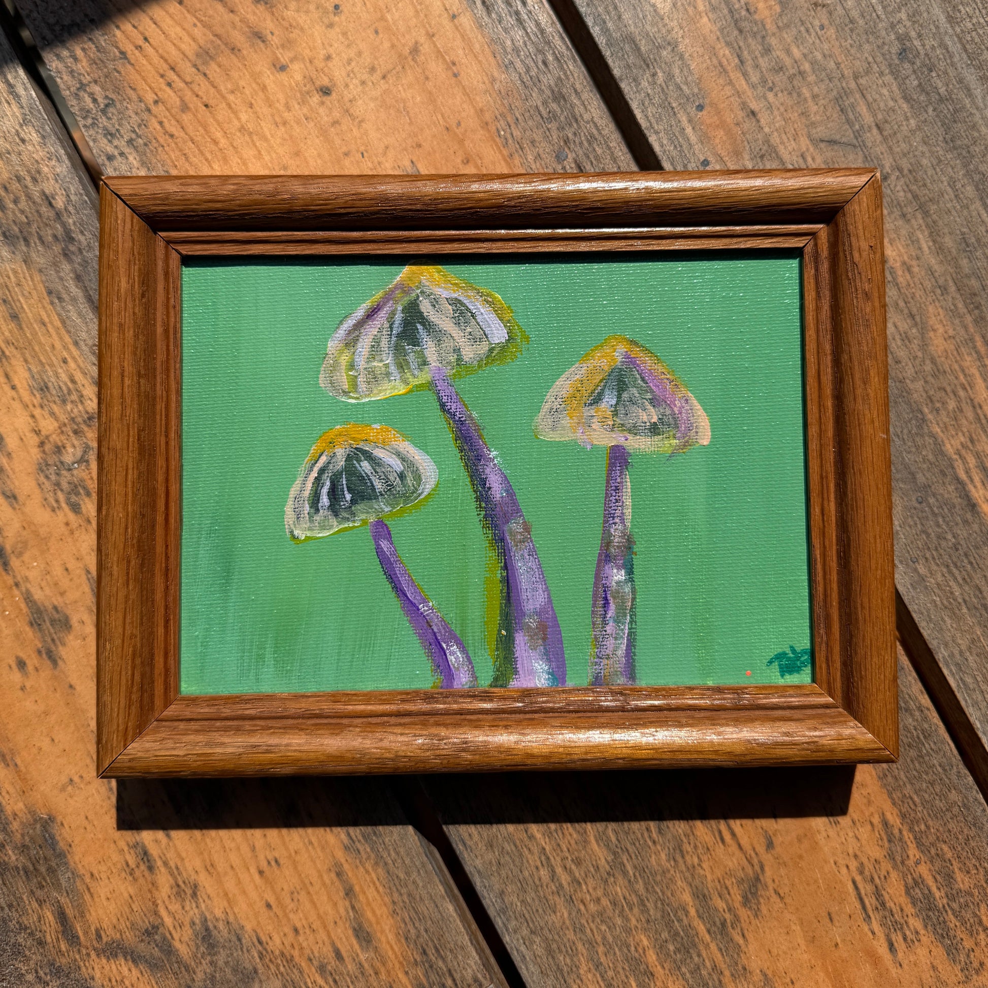 Framed painting of mushrooms with a green background, displayed on a wooden surface.
