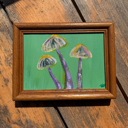 Framed painting of mushrooms with a green background, displayed on a wooden surface.