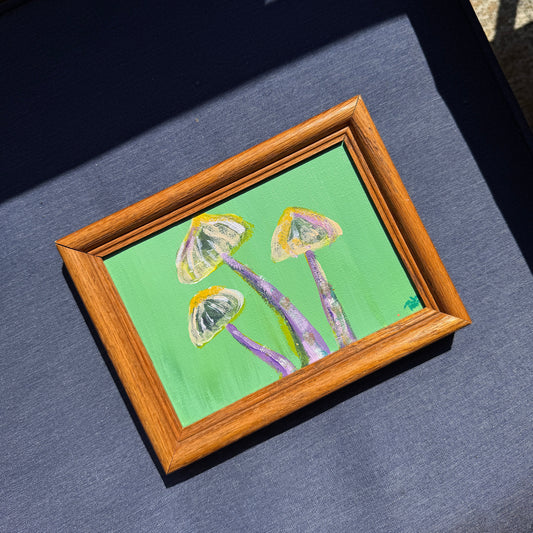Small green and purple painting of mushrooms in a wooden frame on a blue textured surface.