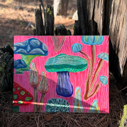 Mixed media artwork, Fun Fungi, features whimsical mushrooms in bright, saturated colors, combining playful design with organic textures. The artwork is photographed in a hollow tree stump in the forest.
