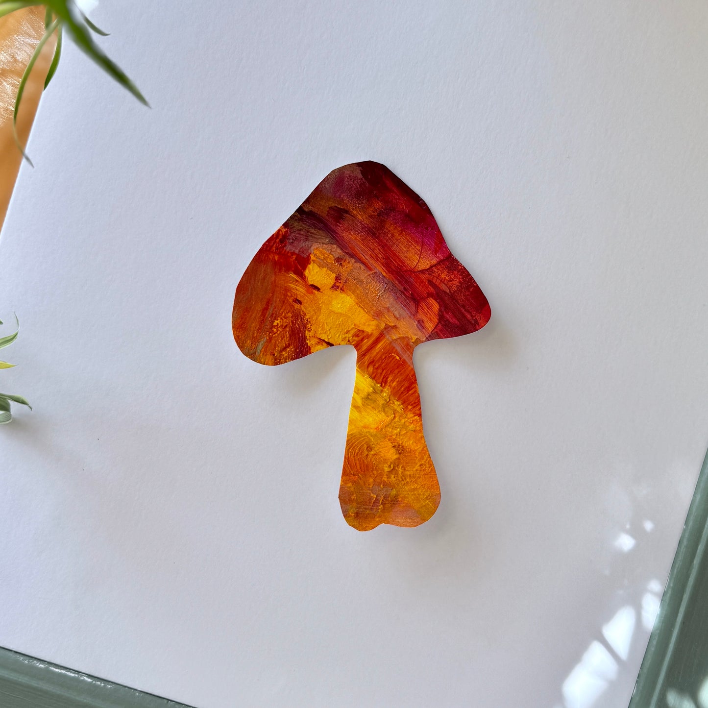 Vibrant, high-quality mushroom art sticker. The sticker is placed on a white surface with plants surrounding the frame, the colors are fiery and vibrant, of orange and red.