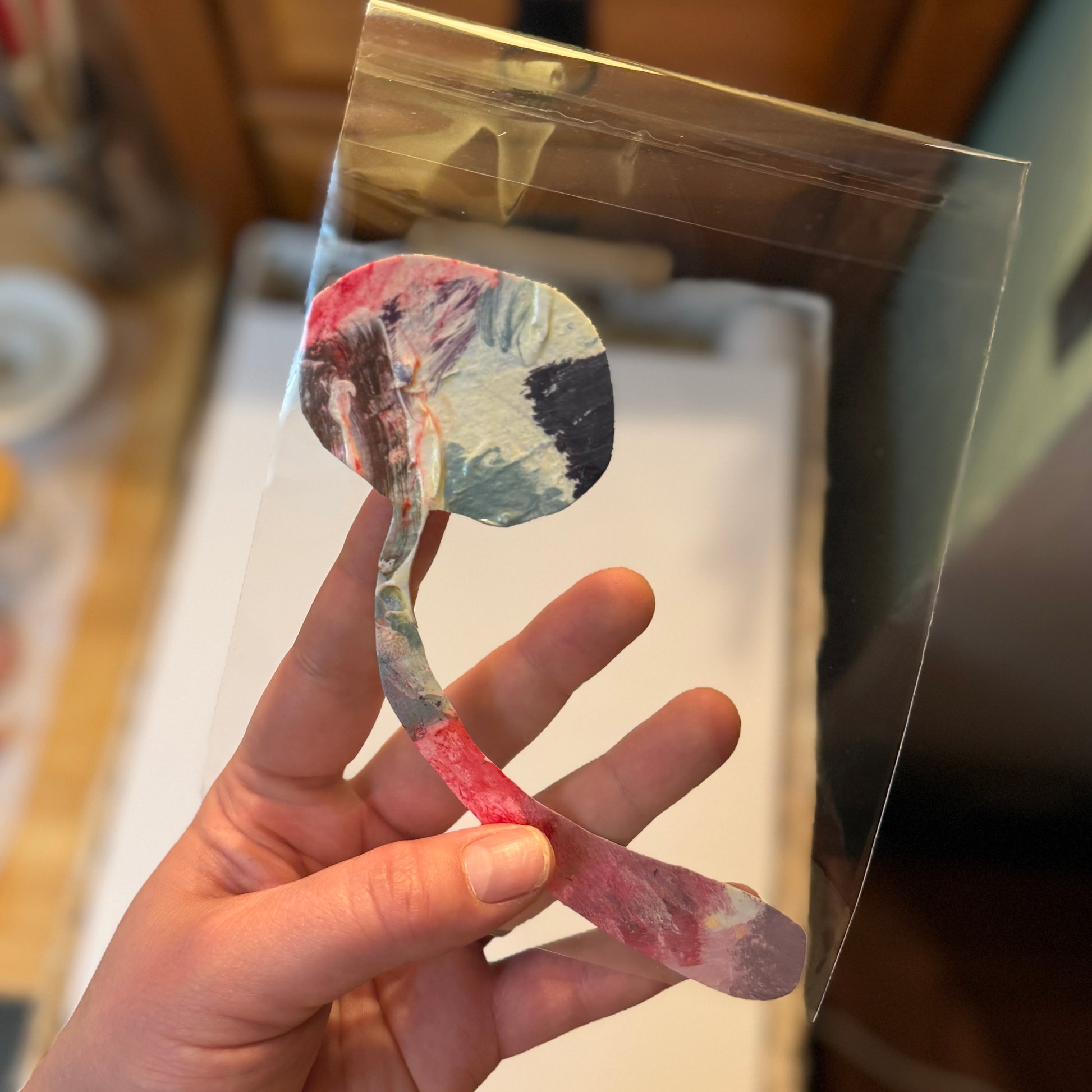 Multicolored, pink mushroom sticker held by the artist. The handmade sticker is in a clear bag and shown in the artist's studio.