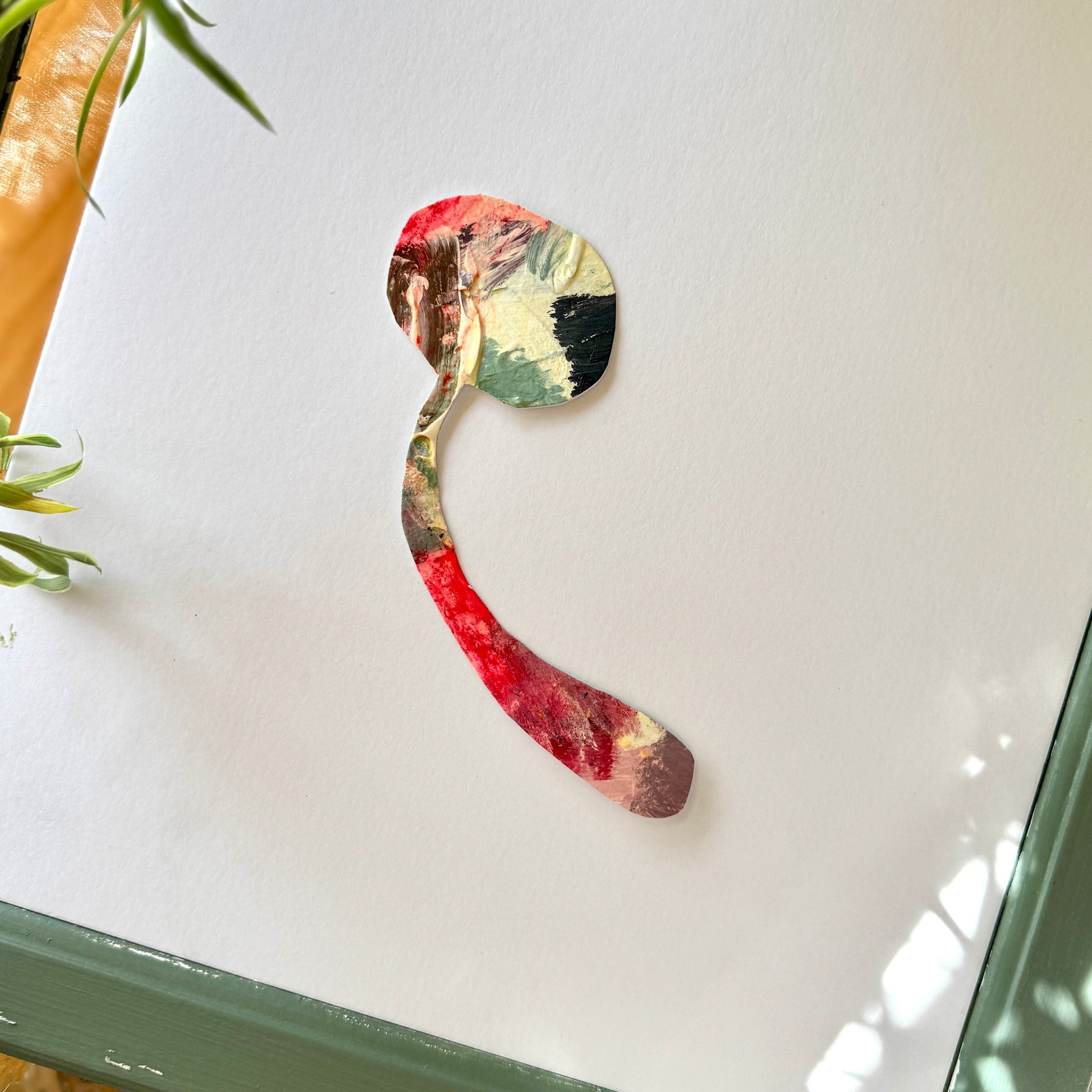 Multicolored, pink mushroom sticker on a white surface with green plants surrounding it. The mushroom is uniquely crafted from the artist's palette.