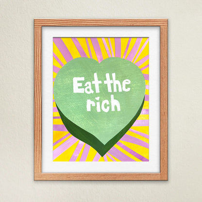 Fine art print hanging on a wall in a white matte and wooden frame. The artwork is bright, colorful, and vibrant. Stripes of bright pink and yellow surround a green candy heart with the typeography “Eat the Rich” in the middle.