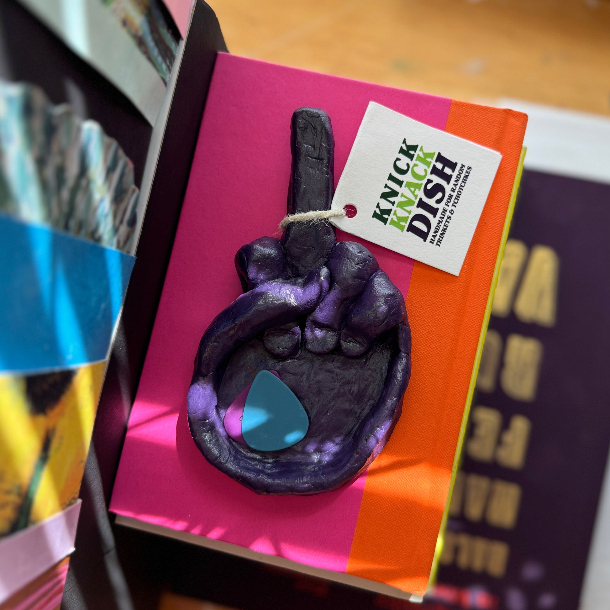 Purple decorative hand dish holding a guitar pick, on a book with 'Knick Knack Dish' tag