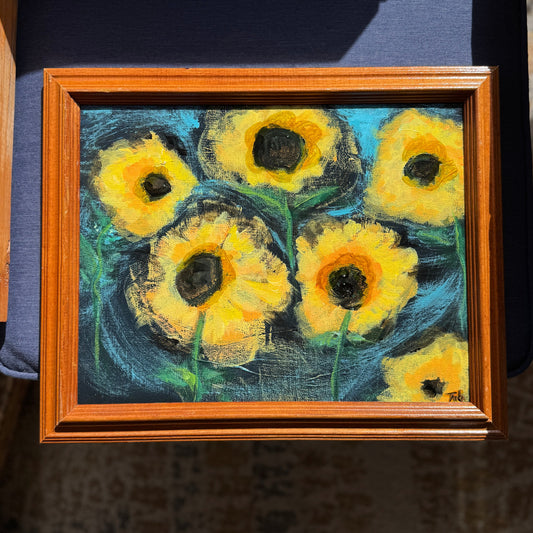 Framed painting of yellow flowers on a dark background outside on a bench.