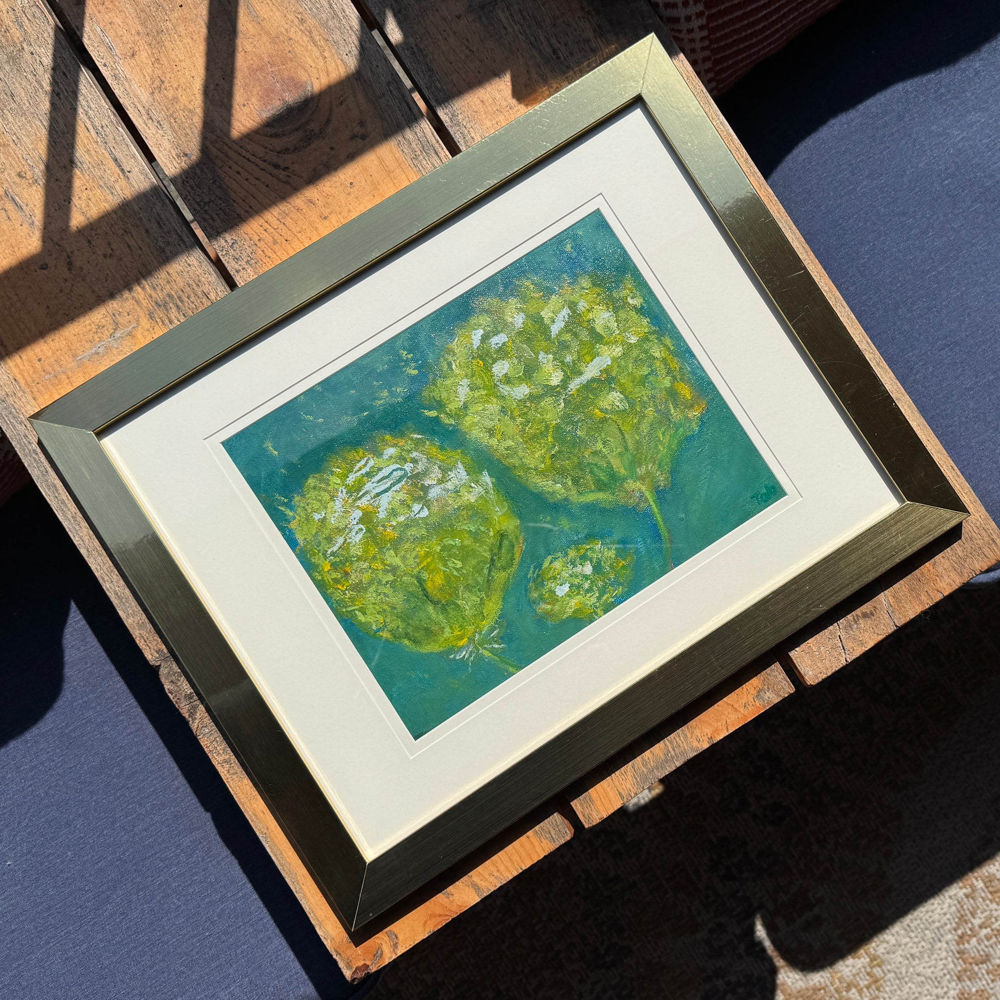 Framed artwork of green abstract fluffy flowers in a golden frame on a wooden bench.