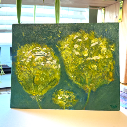 Abstract mixed media painting titled Togetherness, featuring two large yellow flowers with a smaller flower nestled between them. The petals appear to drift and float through the air, set against a painterly green and blue background. The 8x10 canvas artwork evokes connection and harmony with vibrant, expressive brushwork. Shown on a desk. Beautiful light shines through the background.