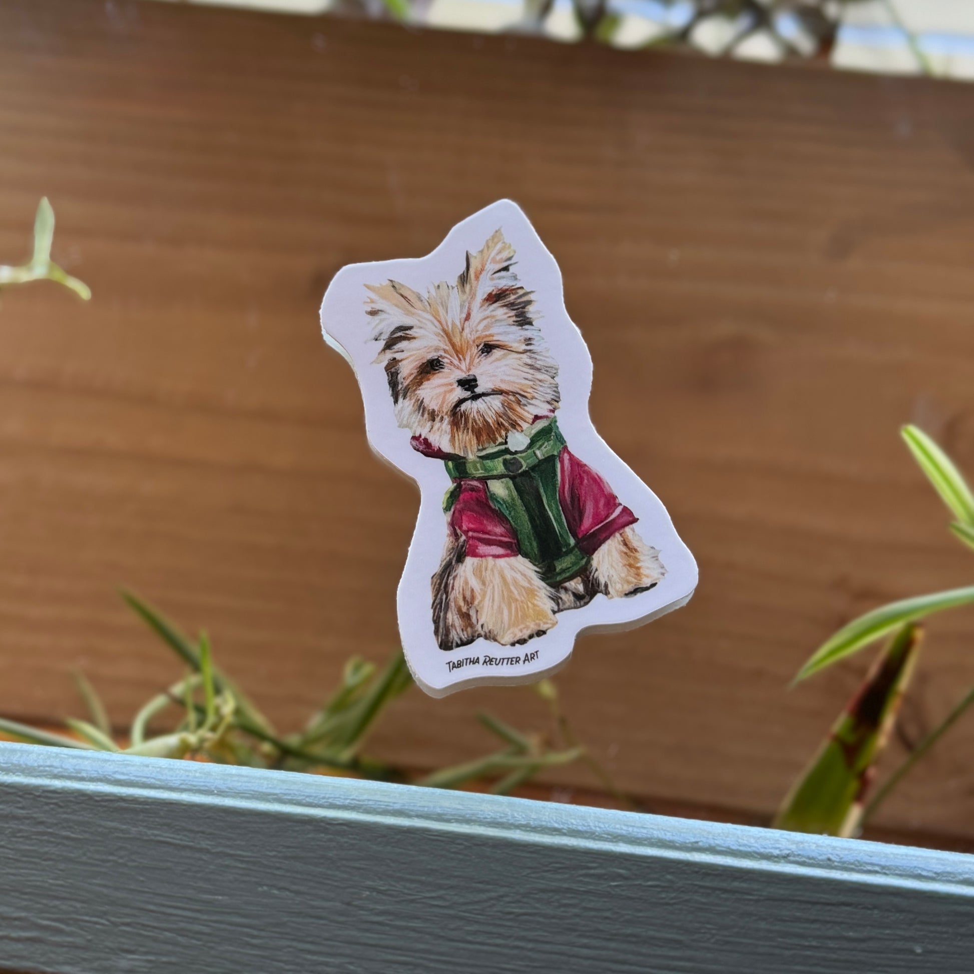 Three-inch vinyl dog sticker on a mirror background. The mirror reflects green plants and a natural wood shelf, adding a natural feel to the image. The sticker is centered, popping against the natural environment. 