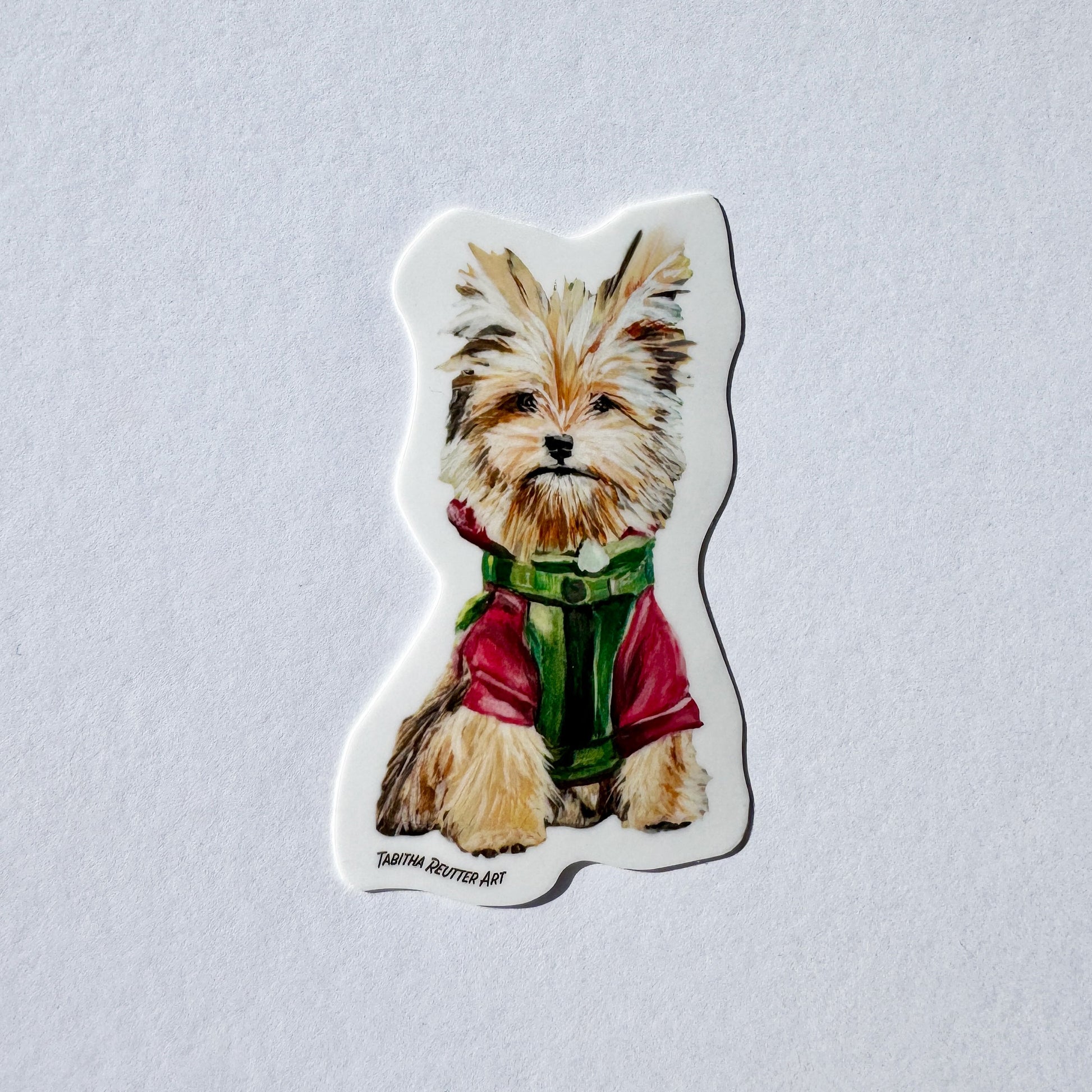 Vinyl sticker of a cute small dog painting. The dog has large ears, light brown fur, and is wearing a red jacket and a green harness. The bottom right corner has the artist’s name, Tabitha Reutter Art.