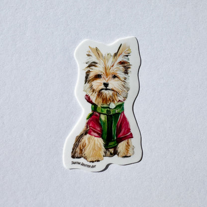 Vinyl sticker of a cute small dog painting. The dog has large ears, light brown fur, and is wearing a red jacket and a green harness. The bottom right corner has the artist’s name, Tabitha Reutter Art.