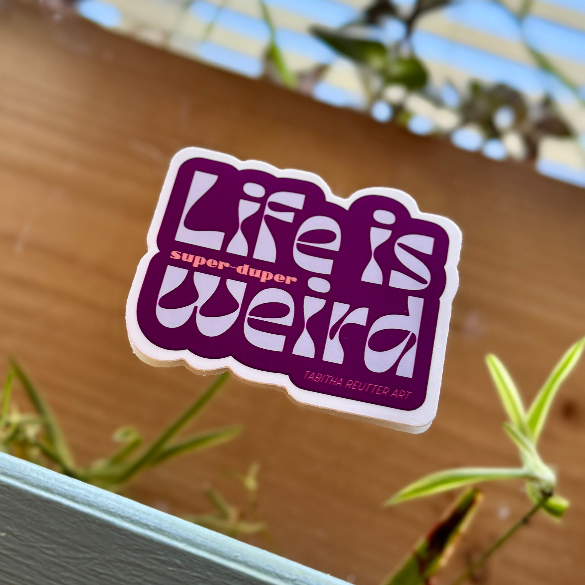 Three-inch vinyl purple sticker features the words “Life is super-duper weird.” The typography is funky and retro, popping colorfully against the background featuring dynamic leafy green spider plants.
