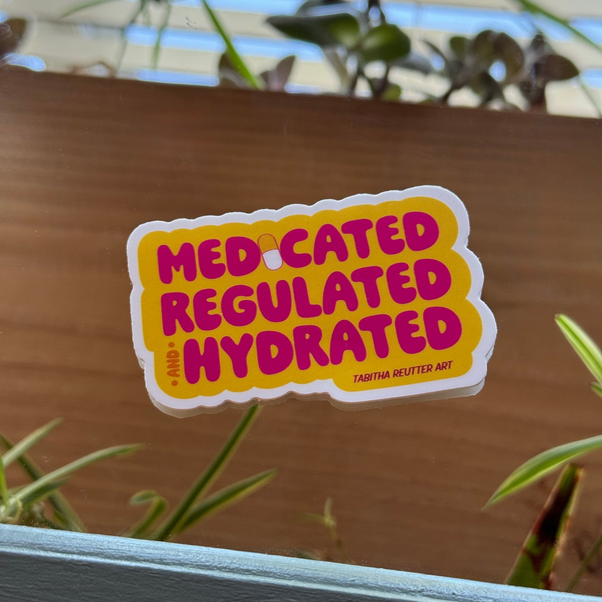 Yellow and pink sticker features the text “medicated, regulated, and hydrated.” The “I” in medicated features an illustration of a pill, while the colors reflect those of a pill bottle. The sticker rests on a mirror, reflecting plants and nature.