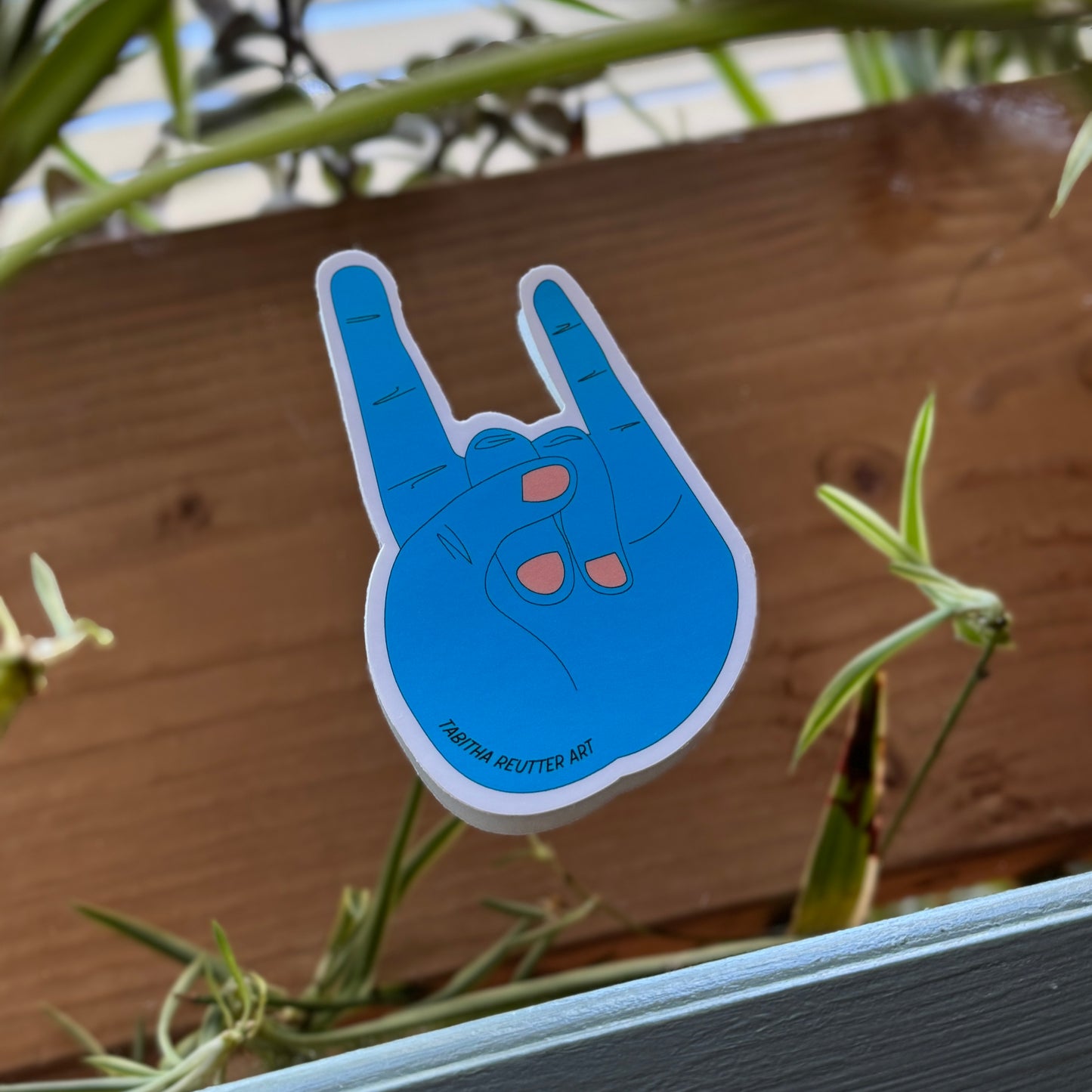 Large, four-inch blue sticker of a rock-on hand and pink painted nails. The sticker is placed on top of a mirror, reflecting plants and wood textures.