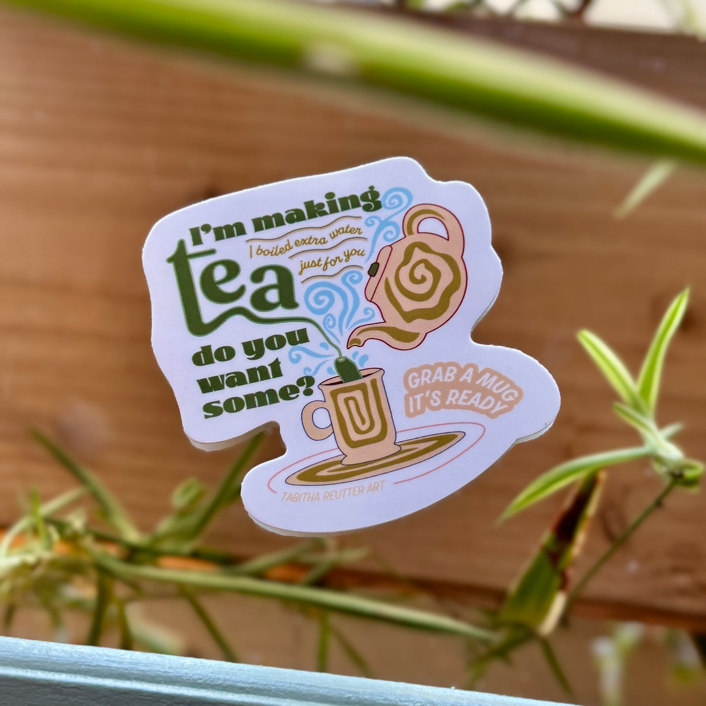 Three-inch vinyl tea sticker on a mirror background. Long, green plants and a natural wood background appear in the background.