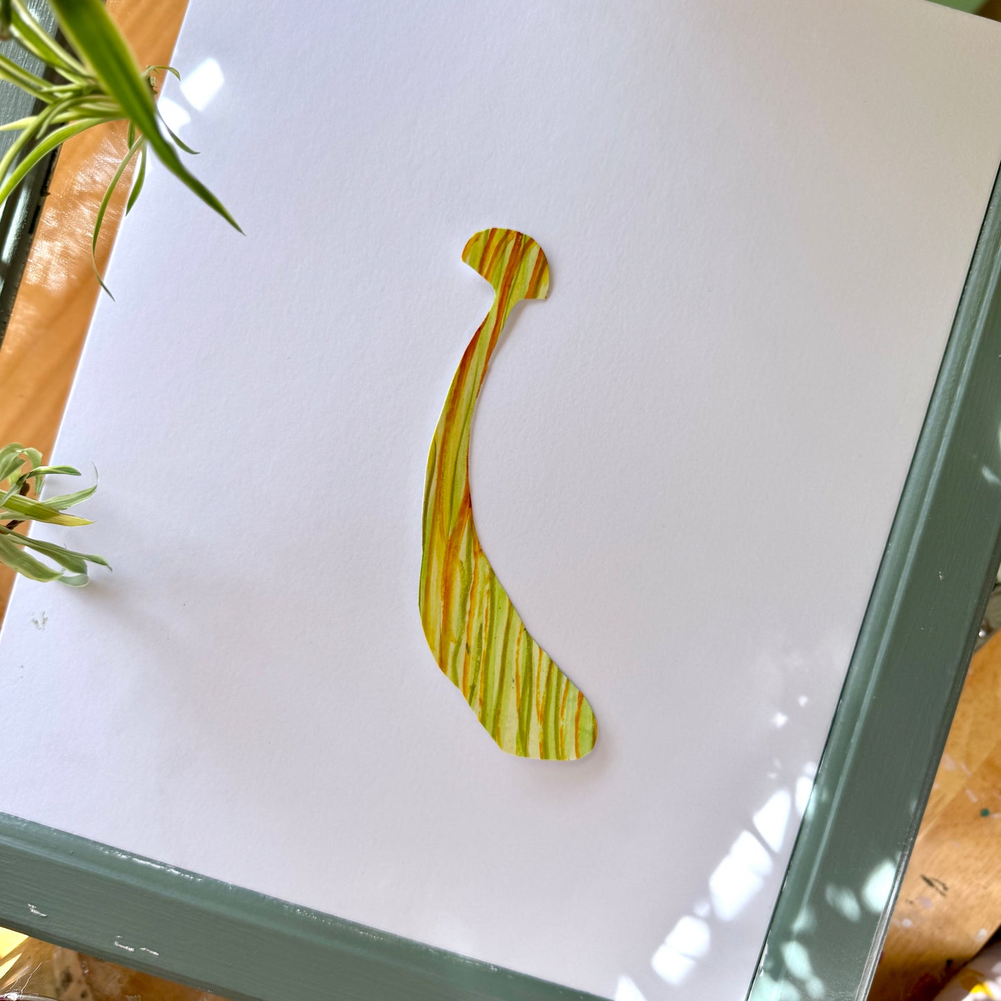 Large, high-quality handmade art sticker. The sticker is yellow with orange and green stripes with a small mushroom cap and a long, thick stem. The sticker is placed on a white surface with plants surrounding the frame.