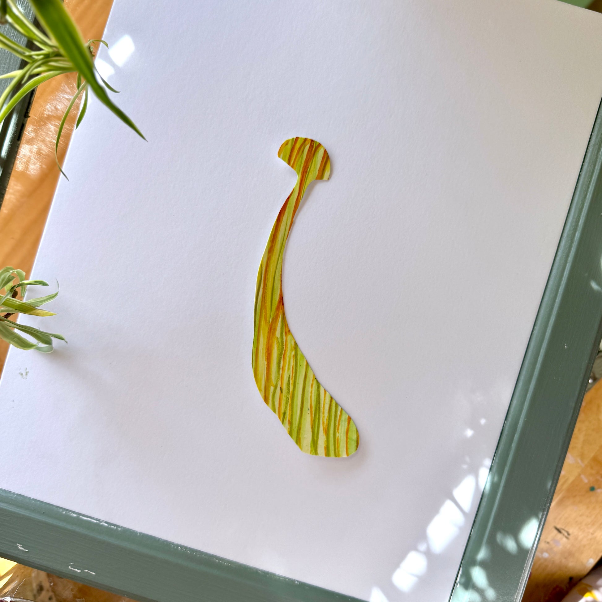 Large, high-quality handmade art sticker. The sticker is yellow with orange and green stripes with a small mushroom cap and a long, thick stem. The sticker is placed on a white surface with plants surrounding the frame.